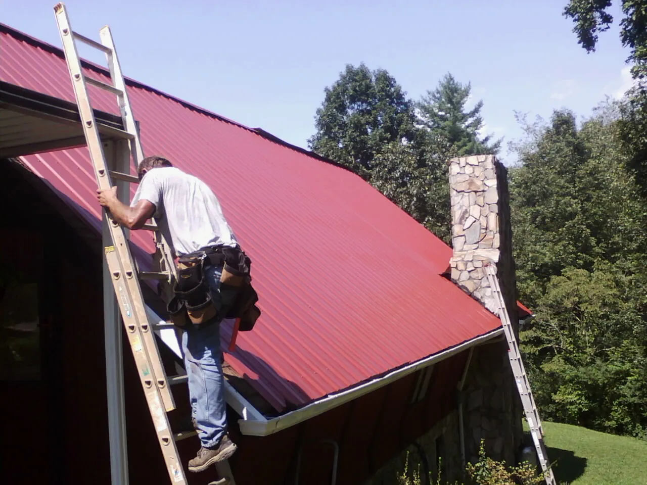 Roofer on a ladder beside a red metal roof and stone chimney — Soffit & Fascia Repair by Roofing Brothers in Eunice